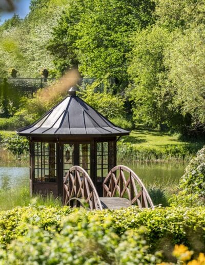 Træpavillon og lille bro ved sø omgivet af grøn beplantning i et klassisk dansk haveanlæg