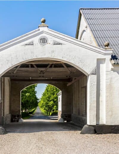 Hvid portbygning fra 1905 med udsigt gennem porten til en lang, træbeklædt allé